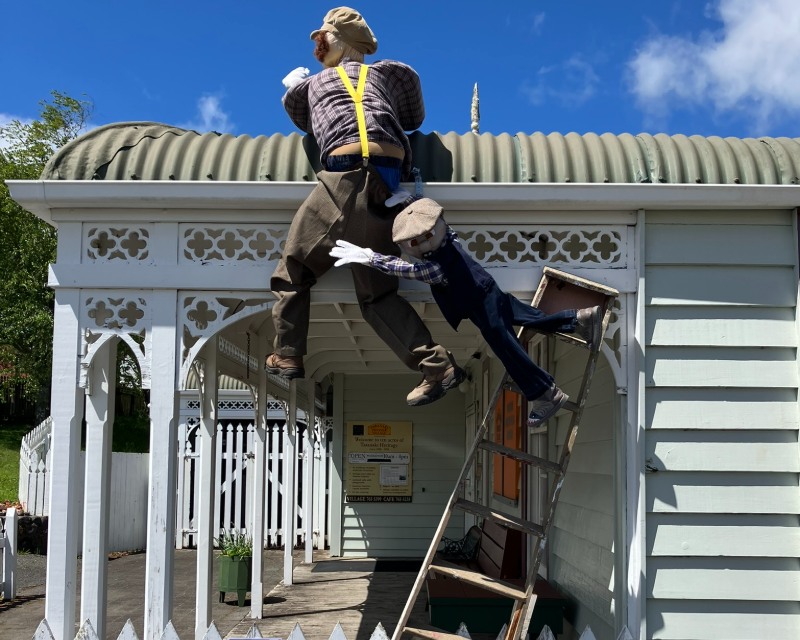 A scarecrow dressed in oldey-timey clothes hangs off the roof of the Pioneer Village, almost falling. A boy scarecrow hangs off the older scarecrows legs, with his own little feet touching on a ladder that's falling.