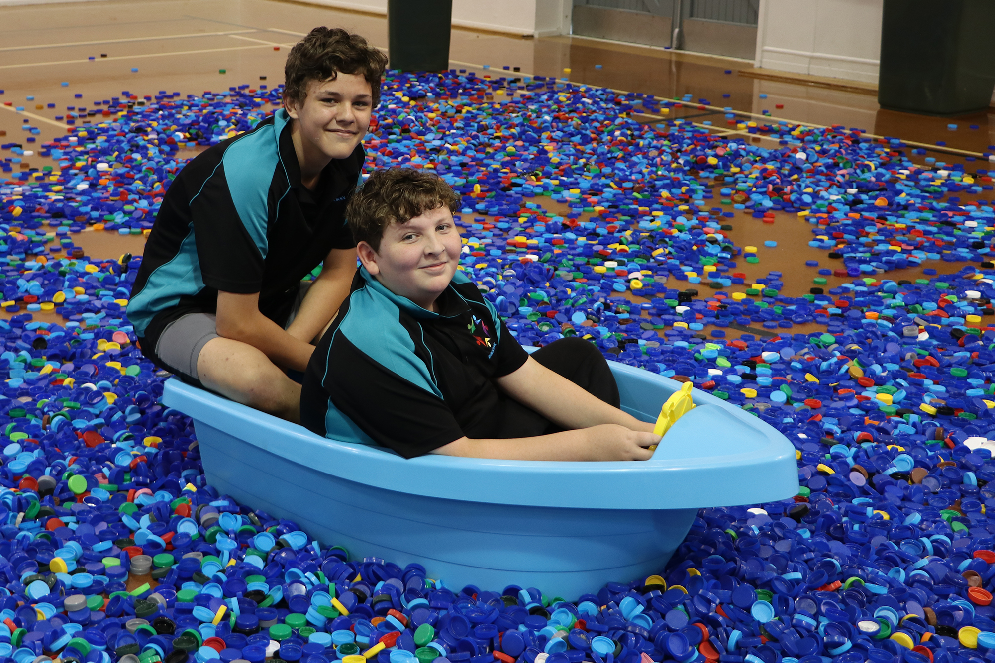 Two students sit in a kids plastic boat in a sea of plastic milk and cream bottle lids.